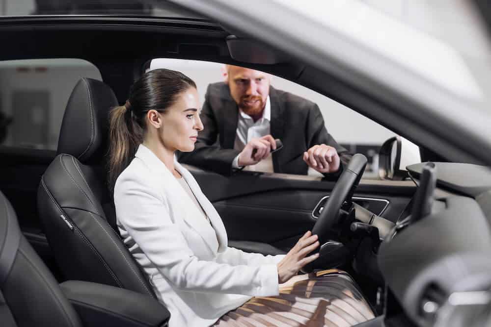 Photo d'une femme derrière un volant de voiture électrique et d'un homme qui lui explique les fonctionnalités de la voiture électrique.